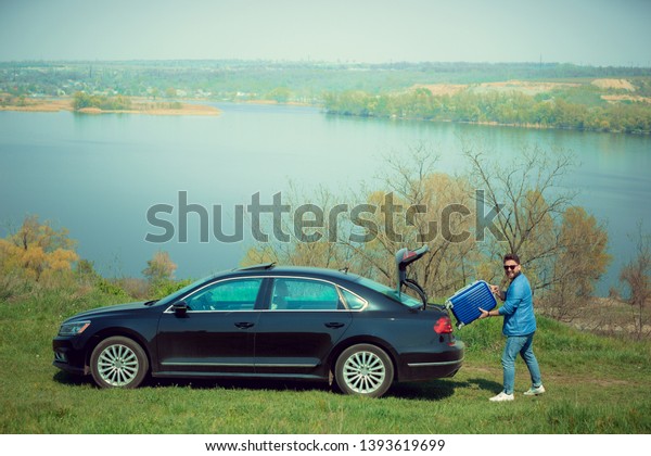 Handsome smiling man in jeans, jacket and\
sunglasses going to vacations, loading his suitcase in car trunk on\
the river\'s side. Preparing for weekend\' trip. Concept of\
summertime, resort,\
chilling.