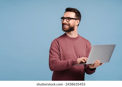 Handsome smiling man, programmer, freelancer holding laptop computer, typing  working online isolated on blue background. Happy confident student studying looking away, copy space. Education concept 