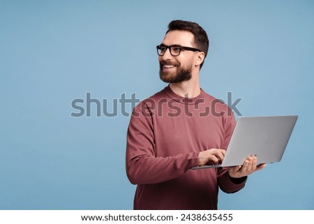 Handsome smiling man, programmer, freelancer holding laptop computer, typing  working online isolated on blue background. Happy confident student studying looking away, copy space. Education concept 