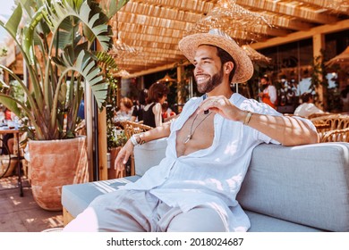 Handsome Smiling Man Sitting At Terrace Restaurant At Summer Time. Stylish Caucasian Man Using Vape Smoke Pen While Wearing Straw Hat And Open Shirt.