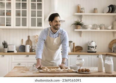 Handsome smiling man wear apron flattening dough for biscuits or home-made dumplings with rolling-pin, stand at dining table, spend time alone in kitchen, enjoy cooking. Hobby, chores, culinary recipe - Powered by Shutterstock