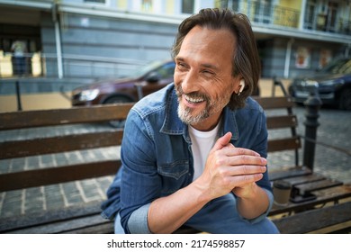 Handsome Smiling Mature Man Spend Time Talking On The Phone Or Listening Music Using Wireless Earphones Sitting On The Bench In Urban City, Enjoying Coffee Wearing Blue Jeans Shirt.