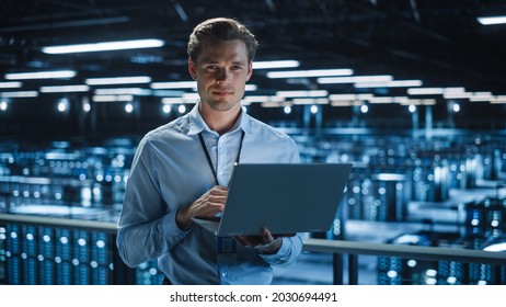 Handsome Smiling on Camera IT Specialist Using Laptop Computer in Data Center. Succesful Businessman and e-Business Entrepreneur Overlooking Server Farm Cloud Computing Facility.