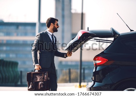 Handsome smiling positive Caucasian bearded businessman in suit with sunglasses holding his briefcase and closing trunk on his car.