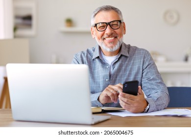 Ein ansprechender lächelnder Senior Mann mit Brille auf dem Handy, während er an seinem gemütlichen Arbeitsplatz mit Laptop zu Hause sitzt, pensionierte männliche Chat mit Freunden in sozialen Medien, tippen auf Smartphone