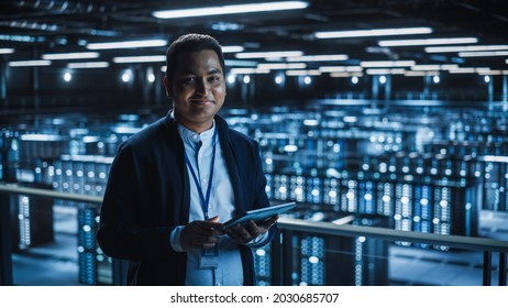 Handsome Smiling IT Specialist Using Tablet Computer In Data Center, Looking At Camera. Succesful Businessman And E-Business Entrepreneur Overlooking Server Farm Cloud Computing Facility.