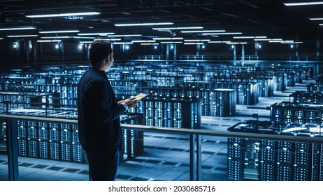 Handsome Smiling IT Specialist Using Tablet Computer in Data Center. Succesful Businessman and e-Business Entrepreneur Overlooking Server Farm Cloud Computing Facility.