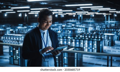 Handsome Smiling IT Specialist Using Tablet Computer In Data Center. Succesful Businessman And E-Business Entrepreneur Overlooking Server Farm Cloud Computing Facility.