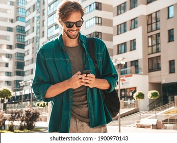 Handsome Smiling Stylish Hipster Lambersexual Model.Man Dressed In T-shirt.Fashion Male Posing In The Street Near White Wall. Holding, Scrolling And Texting In His Cellphone. Writes Sms On Smartphone