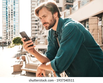 Handsome Smiling Stylish Hipster Lambersexual Model.Man Dressed In T-shirt.Fashion Male Posing In The Street Near. Holding, Scrolling And Texting In His Cellphone. Using Apps