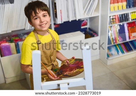 Handsome smiling teenager, talented little artist, holding a palette with oil paints, painting sitting at an easel against the canvases background. Art. Creativity. People. Lifestyle. Hobby. Education