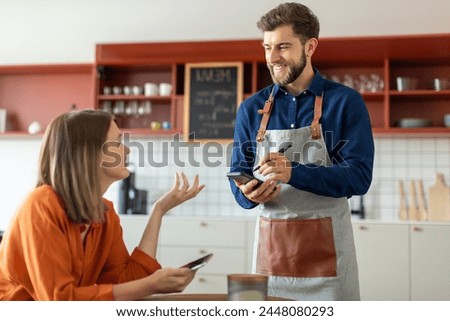 Handsome smiling waiter using application on digital tablet, taking order of young female client in pastry shop of cafe