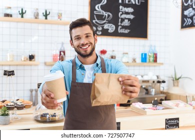 Handsome Smiling Young Barista Holding Coffee To Go In Paper Cup And Take Away Food In Paper Bag