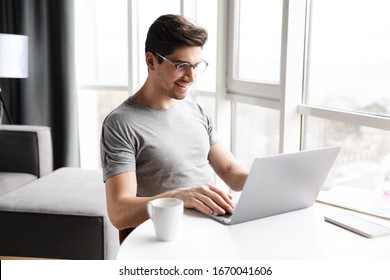 Handsome Smiling Young Bearded Man Wearing Casual Clothes Using Laptop Computer While Sitting At The Table At Home