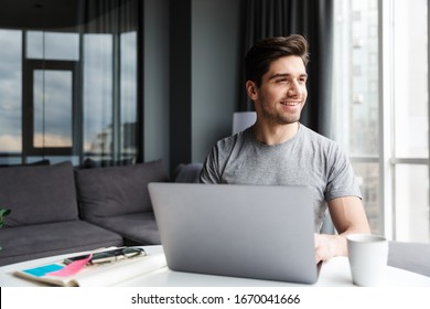 Handsome Smiling Young Bearded Man Wearing Casual Clothes Using Laptop Computer While Sitting At The Table At Home