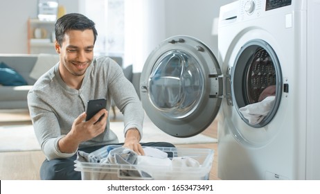 Handsome Smiling Young Man in Grey Jeans and Coat Sits in Front of a Washing Machine and Uses His Smartphone. He Loads Washer with Dirty Laundry. Bright and Spacious Living Room with Modern Interior. - Powered by Shutterstock