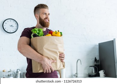 Handsome Smiling Young Man Holding Grocery Bag With Vegetables And Looking Away 