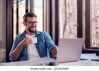 Handsome smiling young man sitting by window in office, drinking coffee while working on laptop. - Powered by Shutterstock