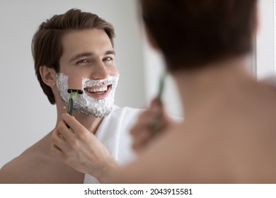 Handsome Smiling Young Man Standing In Bathroom With Towel On Shoulder, Holds Razor Shaving Face Looking In Mirror, Busy In Morning Routine, Caring For Beauty. Healthy Skin, Hygiene, Skincare Concept