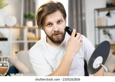 Handsome Smiling Young Man Stands At Home In A White T-shirt And Holds An Electronic Razor While Shaving His Neck, Busy In Morning Routine, Caring For Beauty. Healthy Skin, Hygiene, Skincare Concept