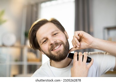 Handsome Smiling Young Man Stands At Home In A White T-shirt And Holds An Electronic Razor While Shaving His Neck, Busy In Morning Routine, Caring For Beauty. Healthy Skin, Hygiene, Skincare Concept