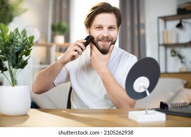 Handsome Smiling Young Man Stands At Home In A White T-shirt And Holds An Electronic Razor While Shaving His Neck, Busy In Morning Routine, Caring For Beauty. Healthy Skin, Hygiene, Skincare Concept