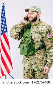 Handsome Soldier In A Cap And With Backpack Talking With His Family Using Mobile Phone After Military Training Near The American Flag In Studio On White Background