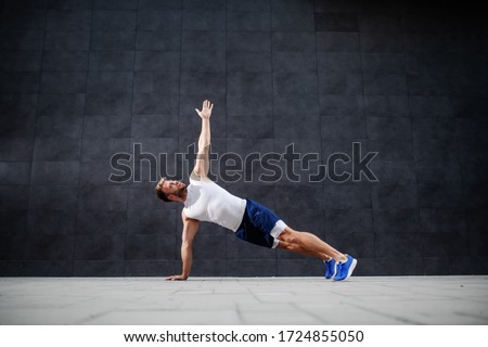 Handsome sporty caucasian man in shorts and t-shirt stretching his arm in plank position in front of gray wall.