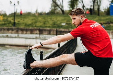 Handsome Sporty Guy Is Streching In A Green Beautiful Park And Is Getting Ready For Jogging While Is Listening Music.