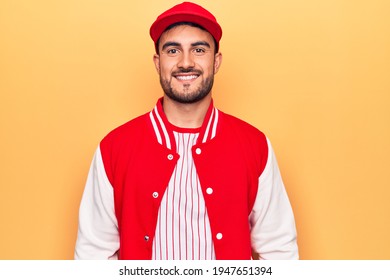 Handsome sporty man with beard wearing baseball jacket and cap over yellow background with a happy and cool smile on face. Lucky person.