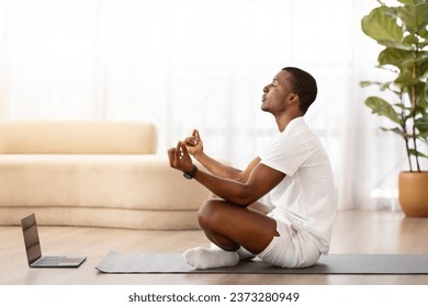 Handsome sporty young black man meditating at home, using laptop, sitting in lotus pose on fitness mat, watching video on Internet, copy space, side view. Virtual yoga classes, stress relief - Powered by Shutterstock