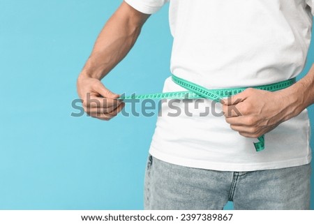 Handsome sporty young man measuring his waist on blue background, closeup. Weight loss concept