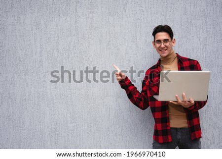 Handsome student with laptop points with gestures to the side. Boy in a red plaid shirt. Exclusive, offer and promotion.