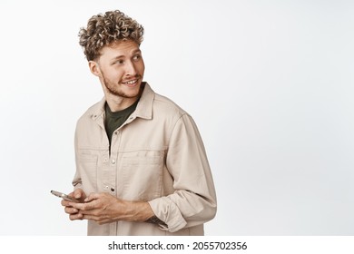 Handsome stylish blond man holding mobile phone, looking behind himself and smiling, reading promo text on copy space, standing against white background
