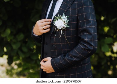 A handsome, stylish groom stands outdoors in a plaid blue suit, a tie with a white rose boutonniere, holding a jacket with his hand. Wedding portrait, photography.