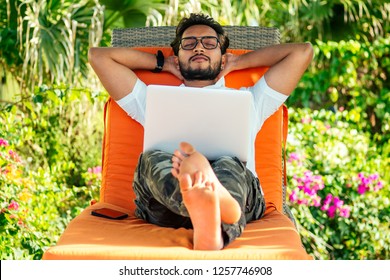 Handsome Stylish Indian Man Summer Vacation In Tropical Paradise Relaxing On An Orange Deckchair Against A Background Of Green Tropical Trees.freelance Remote Work.fitness Male Relaxing On Beach