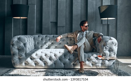 Handsome Stylish Man In Beige Suit At Home Sitting On Sofa.