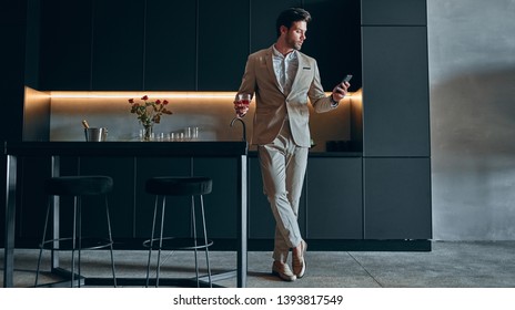 Handsome Stylish Man In Beige Suit At Home. Standing In Modern Black Kitchen With Smart Phone In Hand And Glass Of Whiskey.