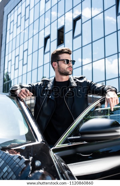 handsome stylish man in leather jacket and\
sunglasses posing near car at\
street