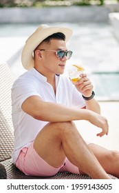 Handsome Stylish Man In Sunglasses And Straw Hat Drinking Old Fashioned Cocktail And Looking At Hotel Swimming Pool