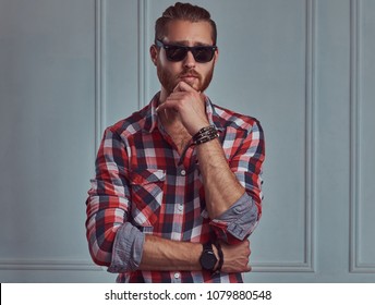 Handsome Stylish Redhead Man In A Flannel Shirt And Sunglasses, Posing In A Studio Against A White Wall.