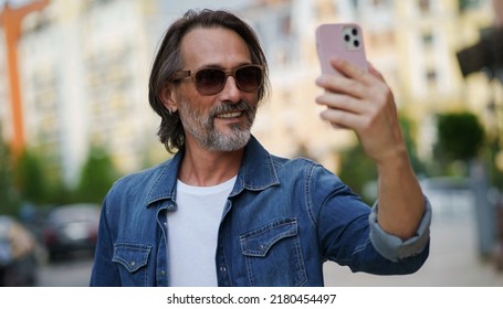 Handsome Stylish Sexy Middle Aged Grey Bearded Man Smiling On Camera Old Town Wearing Casual. Happy Senior Man On Street Of European City Making Selfie Using Mobile Phone. Travel Concept. 