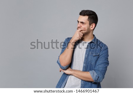 Handsome stylish unshaven young man in denim jeans shirt posing isolated on grey wall background studio portrait. People sincere emotions lifestyle concept. Mock up copy space. Sceptic shocked guy