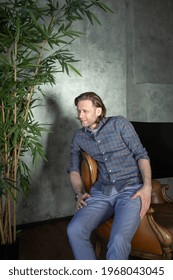 The Handsome Stylish Young Man Sits On An Expensive Leather Armchair Relaxed, Long Curly Hair, He Is Dressed In A Blue Shirt And Trousers, He Is Not Looking At The Camera