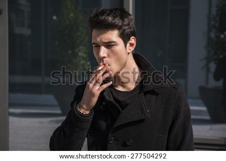 Handsome stylish young man smoking outside in urban setting, looking away