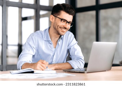 Handsome successful caucasian manager or freelancer wearing glasses, sitting at workplace in office, using laptop, taking notes, studying or working remotely, listening to webinar, smiling - Shutterstock ID 2311823861