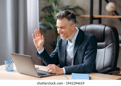 Handsome Successful Confident Mature Business Leader Or Manager, In Formal Suit, Uses Laptop, Communicates Via Video Call, Greetings Colleagues During Online Meeting, Waves His Hand, Smiles Friendly