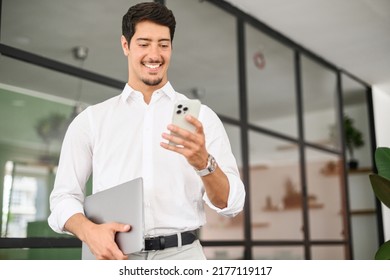 Handsome Successful Hispanic Male Entrepreneur Using Smartphone Indoors, Walking Through Hall And Carrying Laptop, Smiling Businessman Texting On The Phone