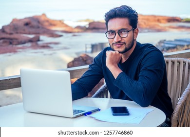handsome and successful indian man in a stylish well-dressed freelancer working with a laptop on the beach.freelance and remote work.businessman student in a summer cafe on the shore of india ocean