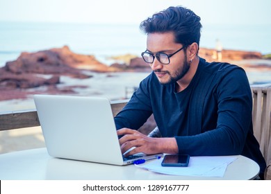 handsome and successful indian man in a stylish well-dressed freelancer working with a laptop on the beach.freelance and remote work.businessman student in a summer cafe on the shore of india ocean